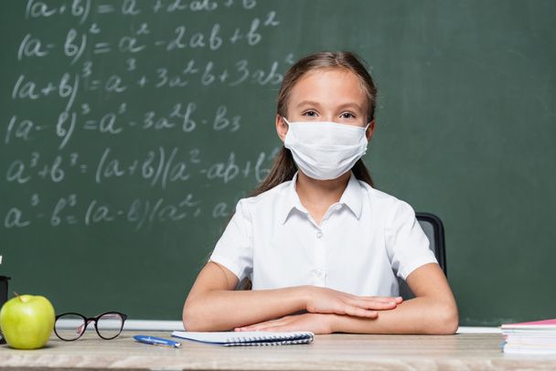 schoolkid in medical mask sitting at desk near apple, notebook, eyeglasses and blurred chalkboard - 写真・画像