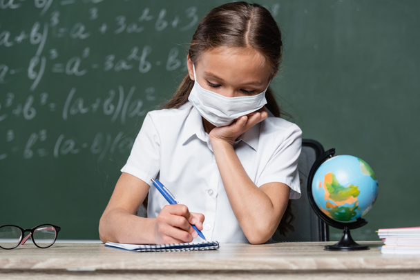 schoolkind in medisch masker schrijven in notebook in de buurt van wereldbol en schoolbord op wazig achtergrond - Foto, afbeelding
