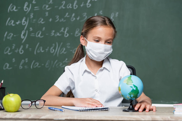 schoolgirl in medical mask looking at globe near notebook, eyeglasses, apple and chalkboard on blurred background - 写真・画像