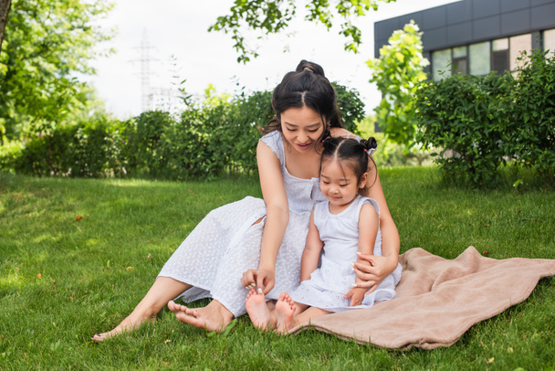 young asian mother touching feet of toddler daughter sitting on picnic blanket  - Foto, Bild