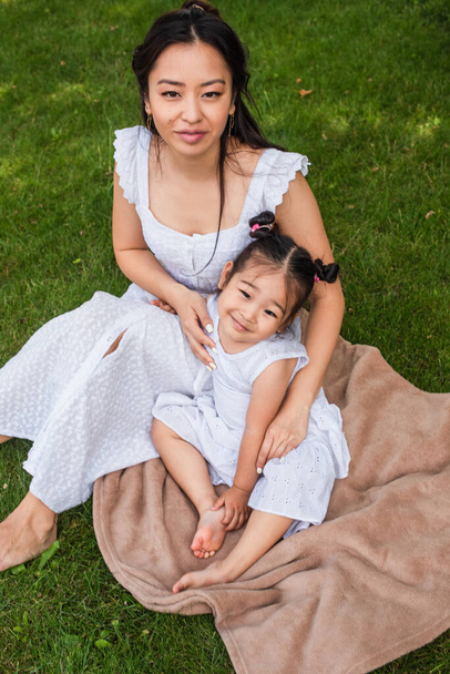 high angle view of asian mother and happy daughter sitting on picnic blanket  - Fotoğraf, Görsel