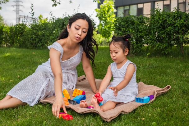 asian mother and toddler girl playing building blocks on picnic blanket in park  - Foto, Bild