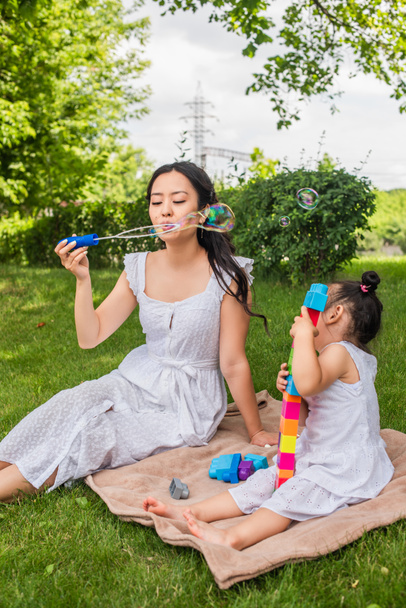 молодая азиатская мать, пускающая мыльные пузыри, пока малышка играет в строительные блоки на одеяле для пикника в парке  - Фото, изображение