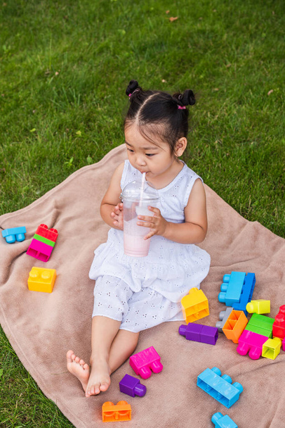 alto ángulo vista de asiático niño niña en vestido beber sabroso batido cerca de bloques de construcción en picnic manta en parque  - Foto, Imagen
