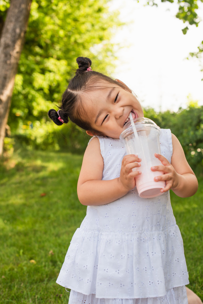 happy asian toddler girl in dress drinking tasty milkshake in park - Photo, Image