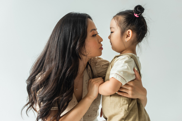 side view of stylish asian mother hugging toddler daughter isolated on grey - 写真・画像