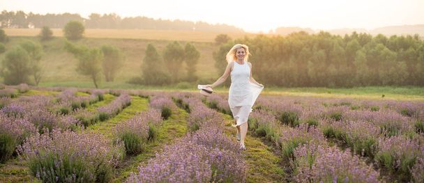 Beautiful young woman wearing a white dress walking in the middle of a lavender field in bloom - Foto, afbeelding