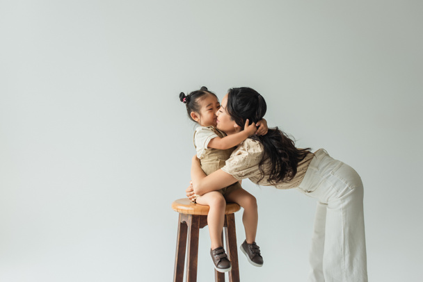 asiático niño chica sentado en silla y besos madre aislado en gris - Foto, Imagen