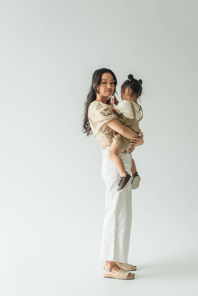 full length of happy asian woman holding in arms toddler daughter on grey - Foto, afbeelding