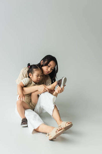 full length of asian mother sitting and wearing shoe of foot of toddler daughter on grey - Фото, изображение