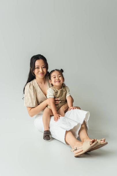 full length of cheerful asian mother sitting with toddler daughter on grey - Zdjęcie, obraz