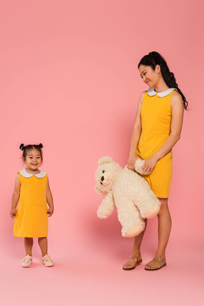 pleased asian woman holding teddy bear and looking at happy toddler daughter on pink - Foto, Imagem