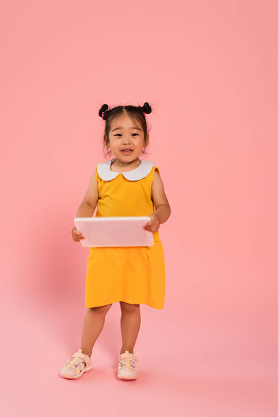 full length of asian toddler kid in yellow dress holding digital tablet on pink - Foto, Imagem
