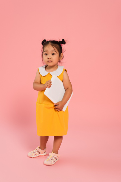 full length of asian toddler girl in yellow dress holding digital tablet on pink - Foto, Bild