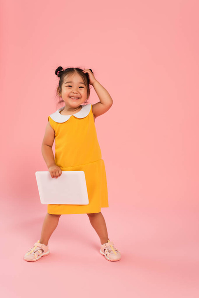 full length of happy asian toddler kid in yellow dress holding digital tablet on pink - 写真・画像