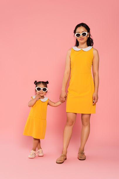 cheerful asian woman holding hands with toddler daughter in sunglasses on pink - Фото, зображення