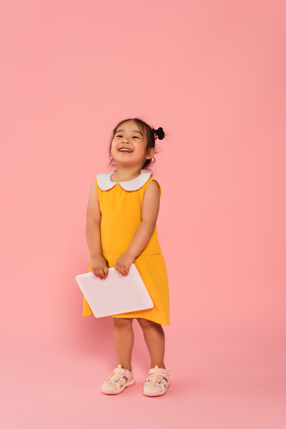 full length of cheerful asian toddler kid in yellow dress holding digital tablet on pink - Foto, Imagem