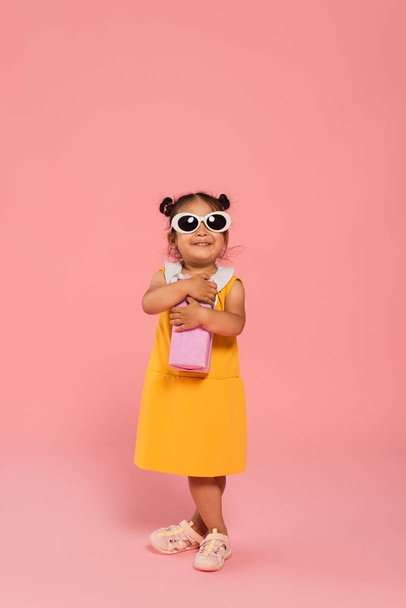 volle Länge der fröhlichen asiatischen Kleinkind in gelbem Kleid und Sonnenbrille hält Geschenk auf rosa - Foto, Bild