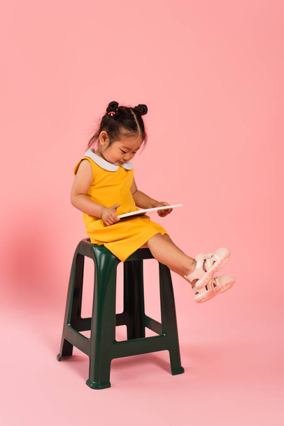full length of pleased asian toddler kid in yellow dress sitting on chair and using digital tablet on pink - Foto, Bild