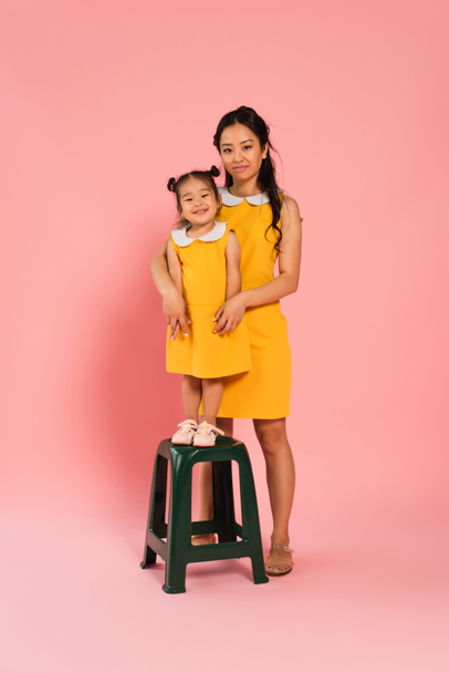 full length of pleased asian toddler kid standing on chair near mother on pink - 写真・画像