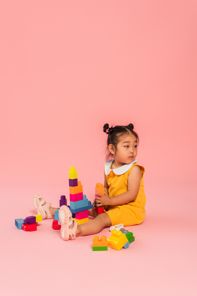 distracted asian toddler girl in yellow dress playing building blocks on pink  - Φωτογραφία, εικόνα