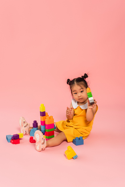 brunette asian girl in yellow dress playing building blocks on pink  - Foto, afbeelding