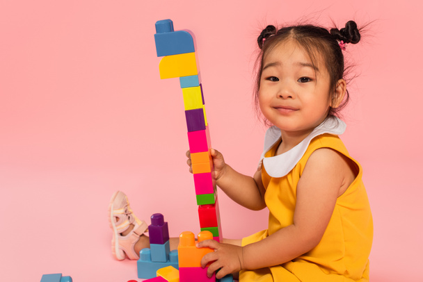 pleased asian toddler girl in yellow dress playing colorful building blocks isolated on pink  - Foto, Imagem