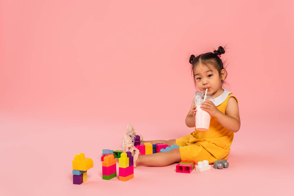 asian toddler kid in yellow dress drinking tasty milkshake through straw near colorful building blocks on pink - 写真・画像