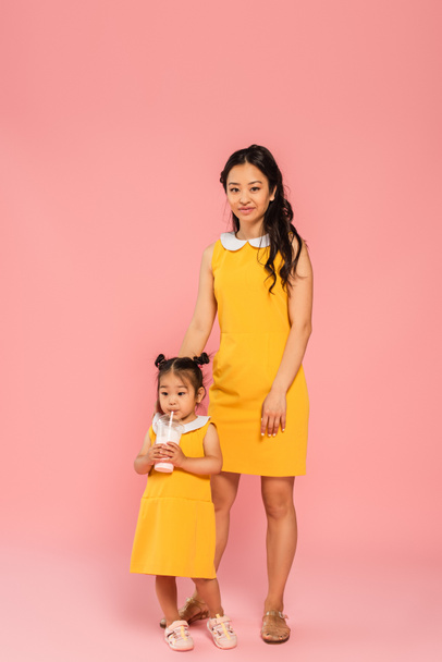 asian toddler kid in yellow dress drinking tasty milkshake through straw near happy mother on pink - Foto, afbeelding