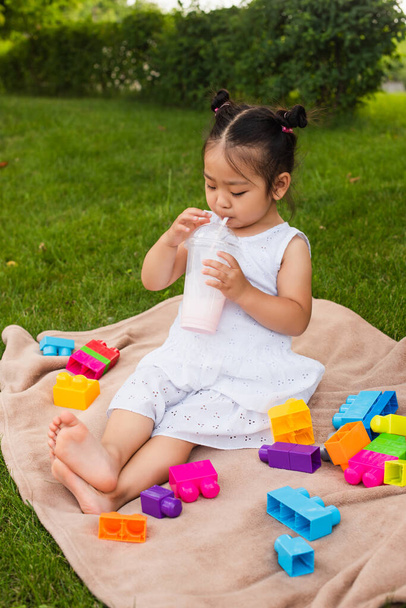 asiatico bambino ragazza in abito bere gustoso milkshake vicino costruzione blocchi su picnic coperta  - Foto, immagini