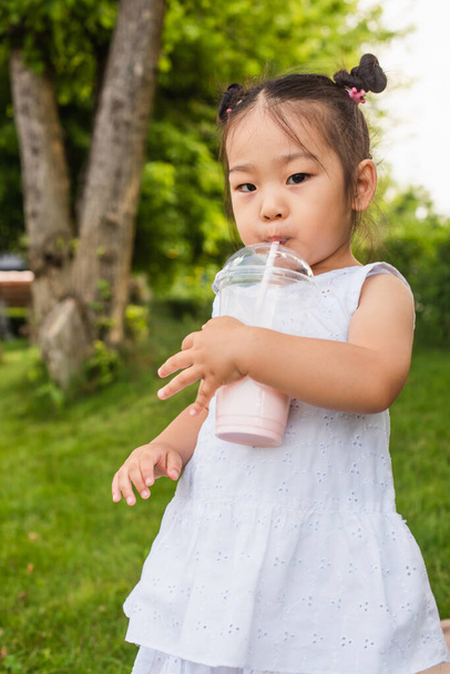 asian toddler kid in dress drinking sweet milkshake through straw in park - Фото, изображение