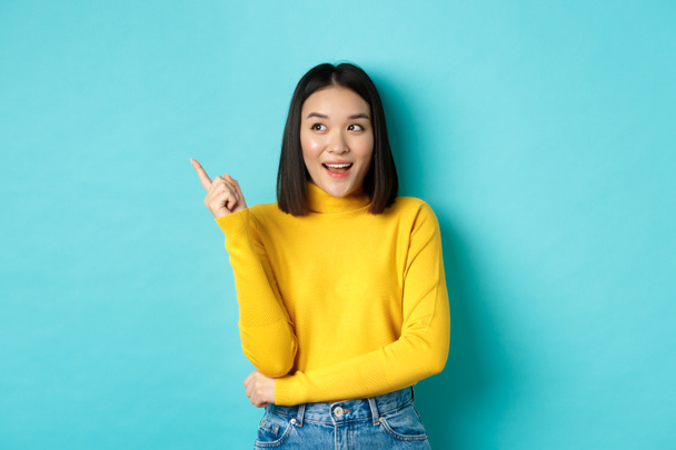 ショッピングコンセプト。黄色のセーターの中に魅力的な韓国の女の子の肖像画,コピースペースにプロモーション提供を示します,ポインティングと幸せな笑顔で左を見ます,青の背景 - 写真・画像