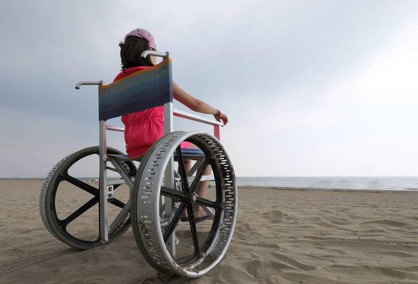 νεαρή κοπέλα στην αναπηρική καρέκλα στην παραλία δίπλα στη θάλασσα και οι ειδικές ρόδες να κινούνται ομαλά στην άμμο - Φωτογραφία, εικόνα