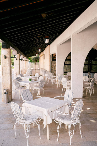 Weiße Tische mit Tischdecken und Stühlen stehen auf der überdachten Veranda des Restaurants mit Säulen - Foto, Bild