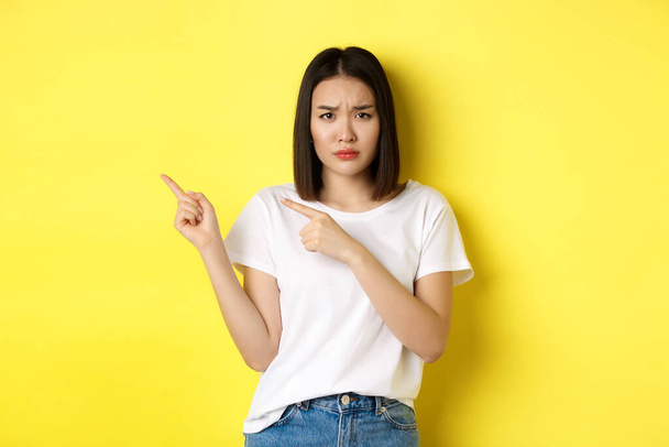 Kauneus ja muoti käsite. Kaunis aasialainen nainen valkoisessa t-paidassa osoittaa sormella vasemmalle, seisoo keltaisen taustan päällä - Valokuva, kuva