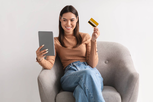 Веселая молодая леди с кредитной картой и планшетным ПК, сидя в кресле, покупки в Интернете, с помощью банковского приложения - Фото, изображение