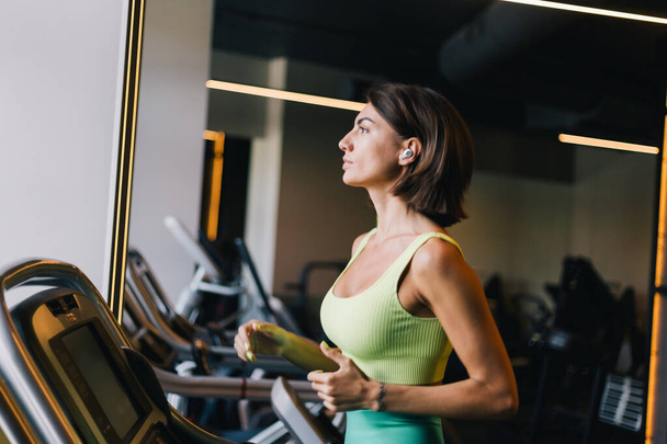 Fit caucasian beautiful woman in fitting sport wear at gym running jogging at treadmill with wireless headphones in ears - Photo, Image