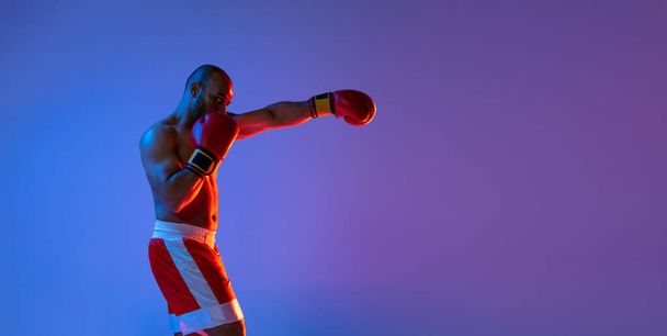 Kırmızı şortlu profesyonel bir boksör, mor arka planda neon ışıkta egzersiz yapıyor. Yan görünüm - Fotoğraf, Görsel