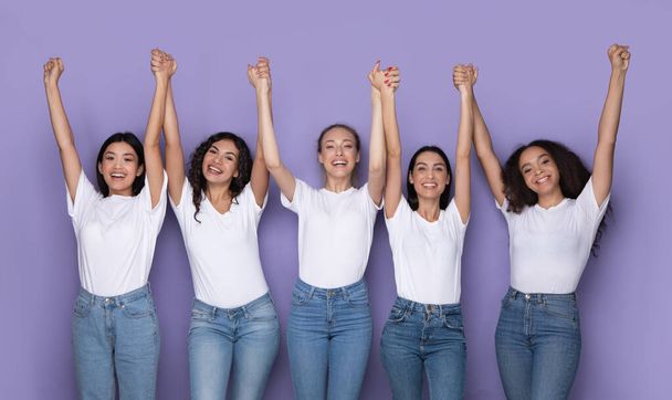 Alegres señoras multiétnicas sosteniendo las manos posando juntas sobre fondo púrpura - Foto, imagen