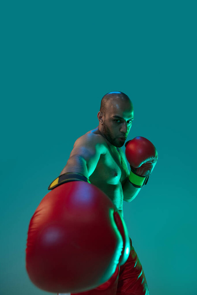 Detailní záběr. Jeden profesionální boxer v červených trenýrkách trénink, cvičení izolované přes modré zelené pozadí v neonovém světle. - Fotografie, Obrázek