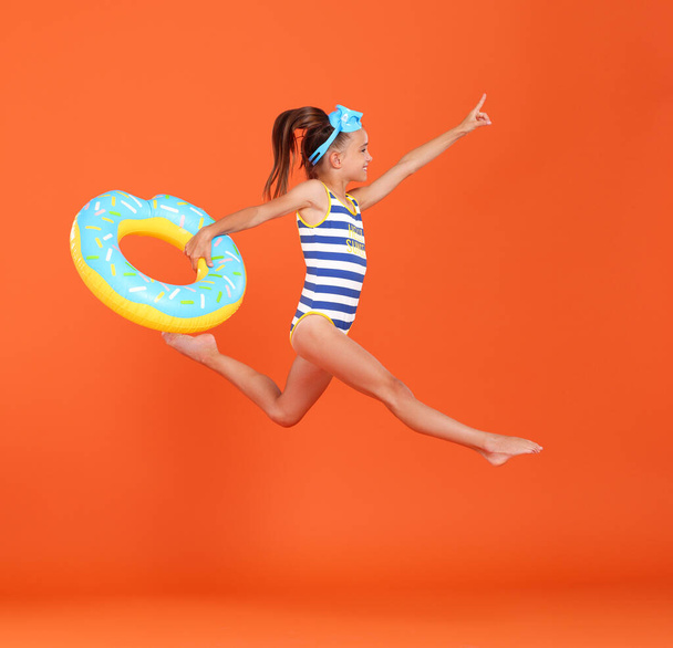 Petite fille mignonne en maillot de bain rayé avec anneau de beignet gonflable à la main, sautant en l'air au ralenti, se dépêche de profiter des vacances d'été en mer, isolé sur fond de studio orange - Photo, image