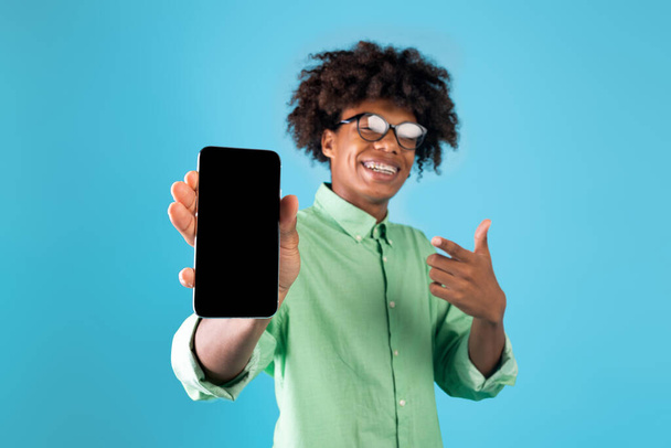 Реклама мобильных приложений. Радостный афроамериканец-подросток показывает на синем фоне чистый экран смартфона - Фото, изображение