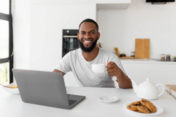 African American man using laptop drinking coffee looking at camera - Photo, Image