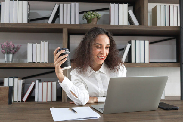 Glücklich lächelnde junge schöne Frau mit lockigem Haar, Kaffeetasse aus Papier und Laptop, weibliches Offizierspersonal mit Konferenzbesprechung online, mobiles Bürokonzept. - Foto, Bild