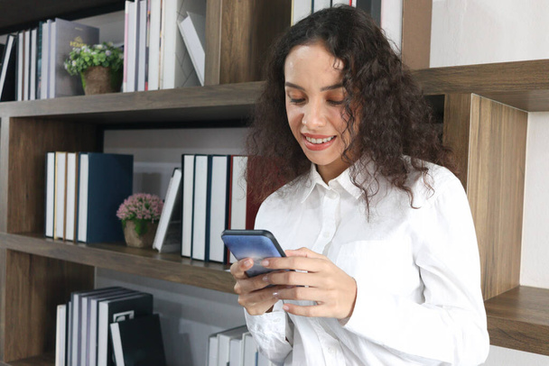 Portrait of happy smiling young beautiful woman with curly hair using mobile phone, female in white shirt texting on smartphones while standing next to the bookshelf in library. - Foto, Imagen