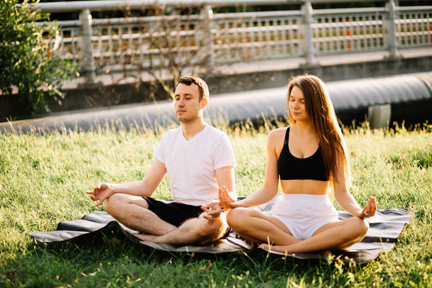 Young couple man and woman meditate while sitting in lotus position, yoga on city lawn, summer evening together - Foto, Bild