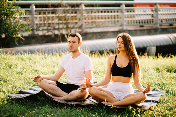 Young couple man and woman meditate while sitting in lotus position, yoga on city lawn, summer evening togetherness - Фото, изображение