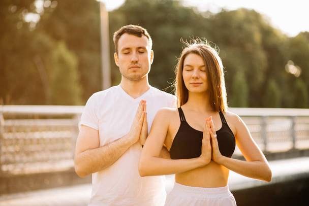 Junges Paar Mann und Frau meditieren gemeinsam im Stehen im Sonnenuntergang, Yoga auf dem Rasen der Stadt, Sommerabend, Lifestyle - Foto, Bild