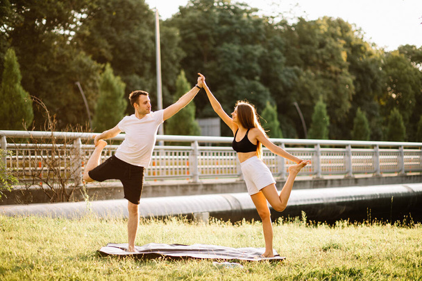 Young couple man and woman doing sports, yoga on city lawn, summer evening, stretching together on sunset - Photo, Image