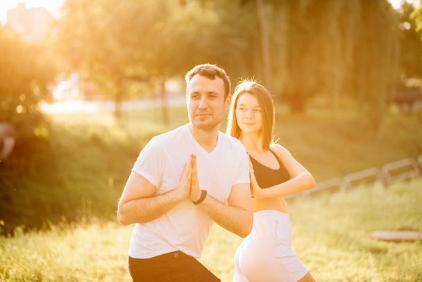 Jovem casal homem e mulher fazendo esportes, ioga no gramado da cidade, noite de verão, alongamento juntos no pôr do sol, concentração - Foto, Imagem